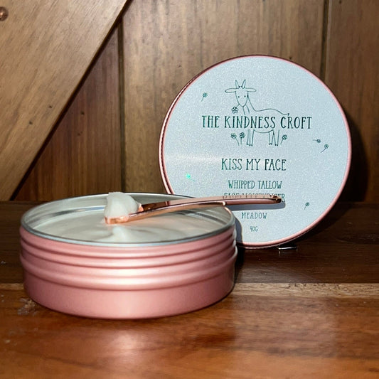 A pink metal tin of whipped facial balm with a small spatula resting inside, shown next to its round lid on a wooden surface. The lid is light blue with black text reading 'THE KINDNESS CROFT KISS MY FACE WHIPPED FALLOW'.