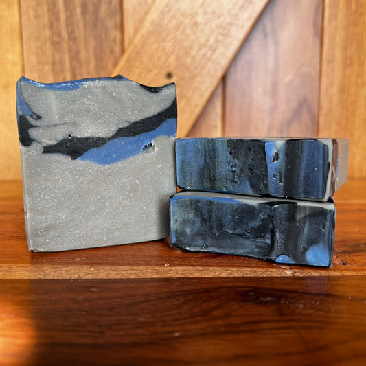 Three handmade soap bars with a marbled slate gray, black, and blue pattern, displayed on a wooden surface against a wooden background.